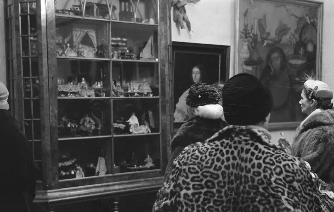 Visitors in winter coats studying framed paintings and a cased dolls’ house at an auction viewing day in 1960