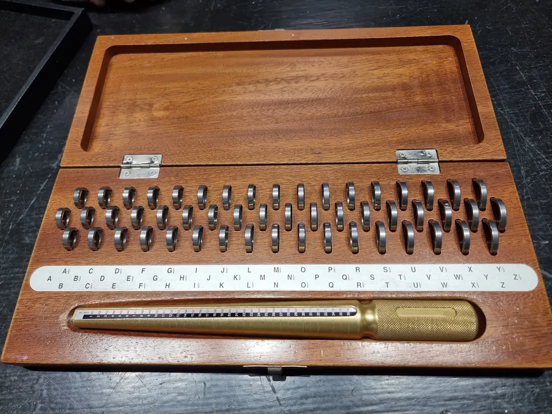 An open wooden case holding rows of steel sizing rings, a strip printed with UK letter sizes, and a brass ring mandrel